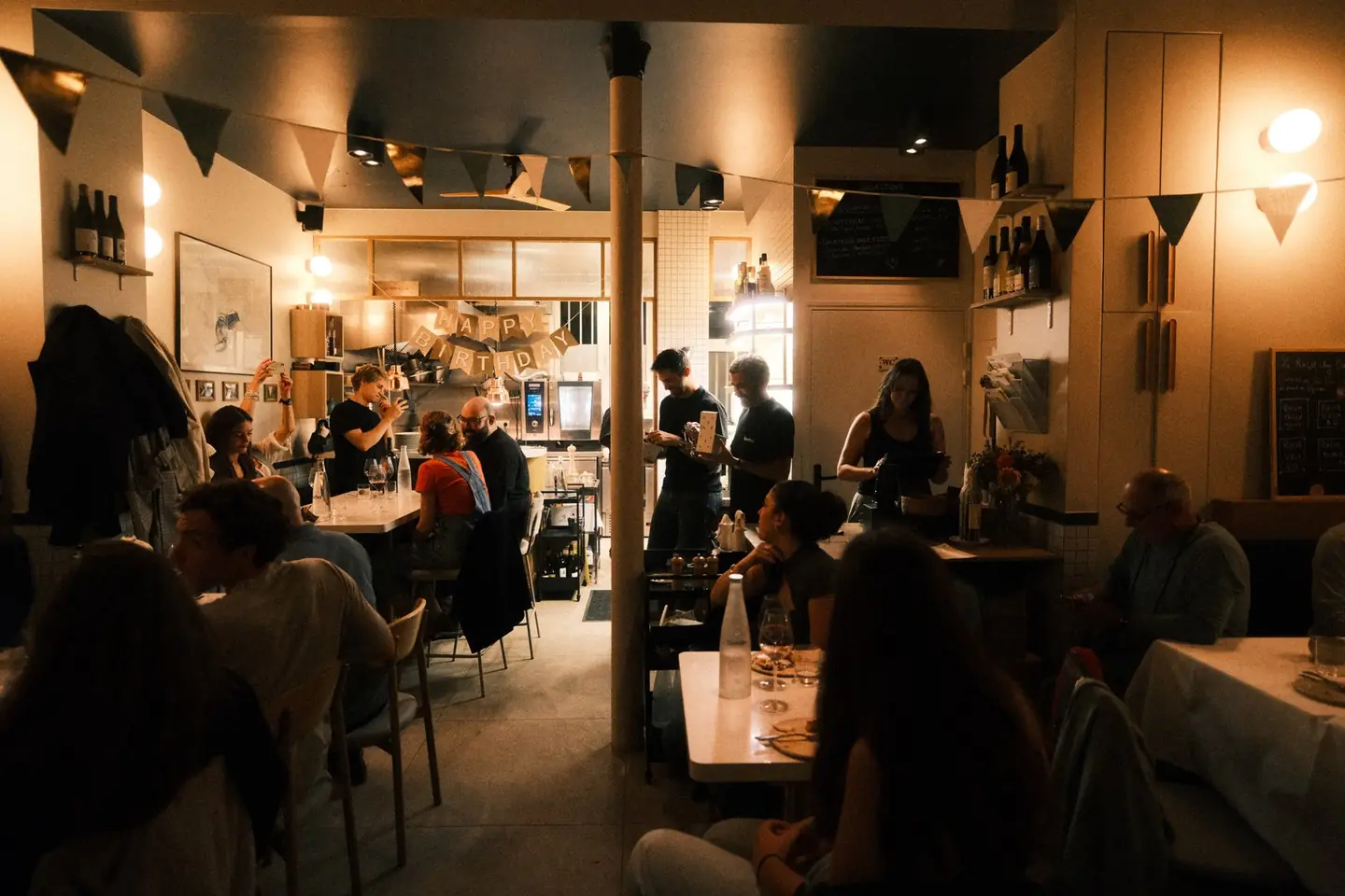 La salle de Barbo pendant le service du soir : tables occupées, équipe en mouvement, cuisine ouverte au fond.